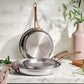 Anolon EverLast stainless steel skillet set with bronze handles on a marble kitchen benchtop, the 25cm pan's base showing the Anolon logo